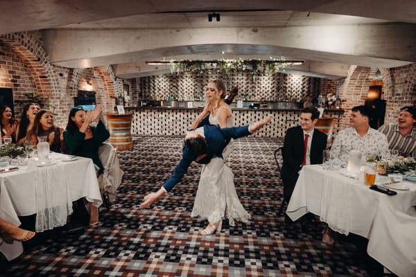 Courtney and Cameron dance on the reception stage at Sandstone Point Hotel — Cellar, surrounded by seated guests applauding and smiling.