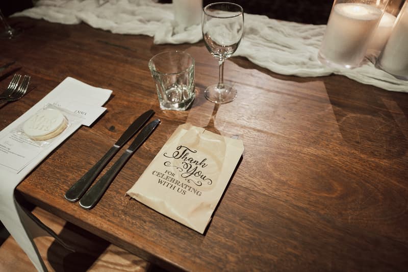 A wooden table at Sandstone Point Hotel — The Cellar reception is set with a white napkin, two knives, a water glass, a wine glass, a cookie with 'Mr & Mrs' written on it, and a paper bag that reads 'Thank You For Celebrating With Us'.