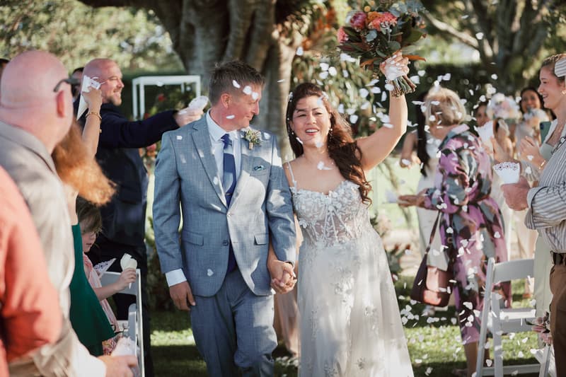 Bride Jheng and groom Dane walk down the aisle at Sandstone Point Hotel — The Pavilion, holding hands as guests throw white confetti.