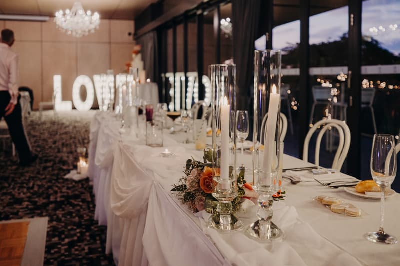 Long reception table set with white linens, glassware, candles in tall holders, floral arrangements, and bread rolls at Sandstone Point Hotel — Pumicestone Room, with illuminated 'LOVE' letters in the background and a guest walking nearby.