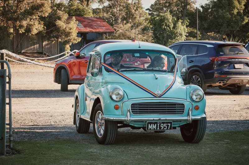 A vintage light blue car decorated with a rainbow ribbon drives on a gravel path at Sandstone Point Hotel — The Pavilion, with two people visible inside the car.