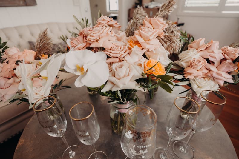 Wedding floral arrangements with pink and white roses and orchids in glass jars on a table at Preston Peak Winery Functions Centre, accompanied by champagne flutes with gold rims.