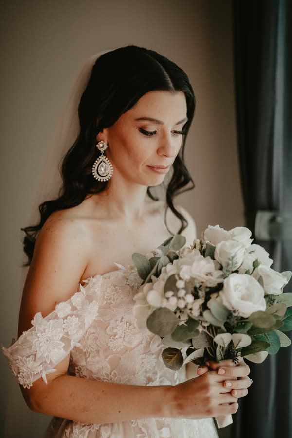 Bride in an off-shoulder lace wedding gown holding a bouquet of white flowers and greenery, standing indoors near a window with a curtain.