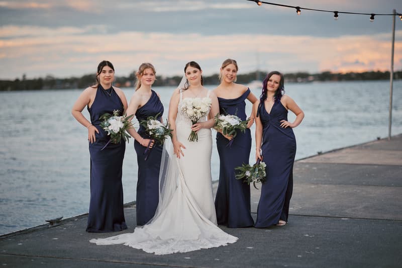 The bride Terion stands with four bridesmaids dressed in navy blue gowns holding bouquets at Sandstone Point Hotel, posing on a waterfront pier during the couple portraits stage.
