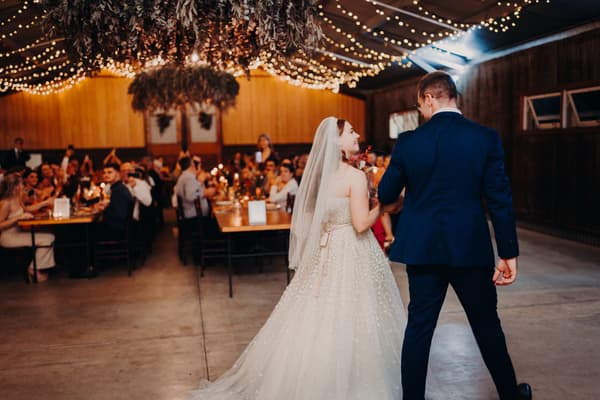 Bride Lilly and groom Connor walk together at the reception stage at Yabbaloumba Retreat — The Shed, with guests seated at tables decorated with candles and string lights overhead.