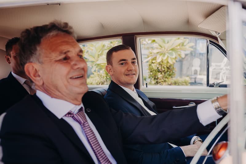 The groom Dylan sits in the back seat of a car, looking toward the camera, while an older man, possibly the groom's father, sits in the front passenger seat smiling. Another man is partially visible in the back seat behind them.