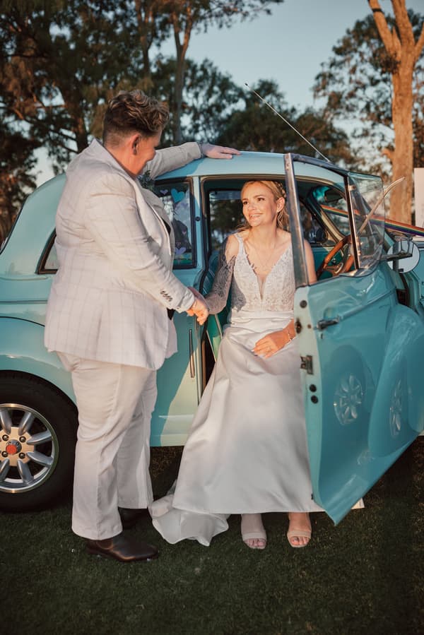 Bride Mandy sits inside a vintage light blue car with the door open, holding hands with bride Selina who stands outside the car at Sandstone Point Hotel.