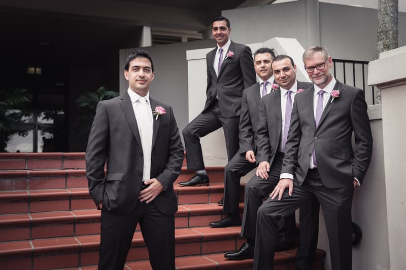 The groom and four groomsmen pose on the steps at Hillstone St Lucia — The Quartyard, all wearing dark suits with pink boutonnieres and lavender ties except the groom who has a white tie.