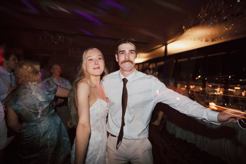 The bride Morgan and the groom Joel pose together on the reception stage at Sandstone Point Hotel — The Pumicestone Room, with guests dancing in the background.