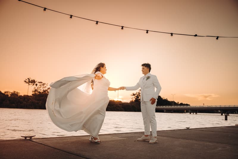 Brooke and Tiffany hold hands near the water at sunset, with Brooke's wedding dress flowing in the breeze at Sandstone Point Hotel.
