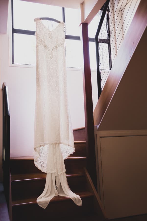 The bride's wedding dress is hanging on a hanger in front of a window at Toowong Rowing Club, displayed above a wooden staircase.