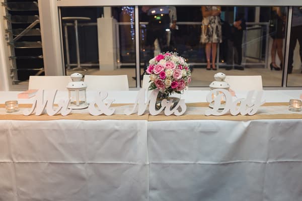 Reception table at Bilinga Beach Weddings — Bilinga SLSC decorated with a white tablecloth, a burlap runner, white lanterns, candles, a bouquet of pink and white roses, and white wooden letters spelling 'Mr & Mrs Day'.