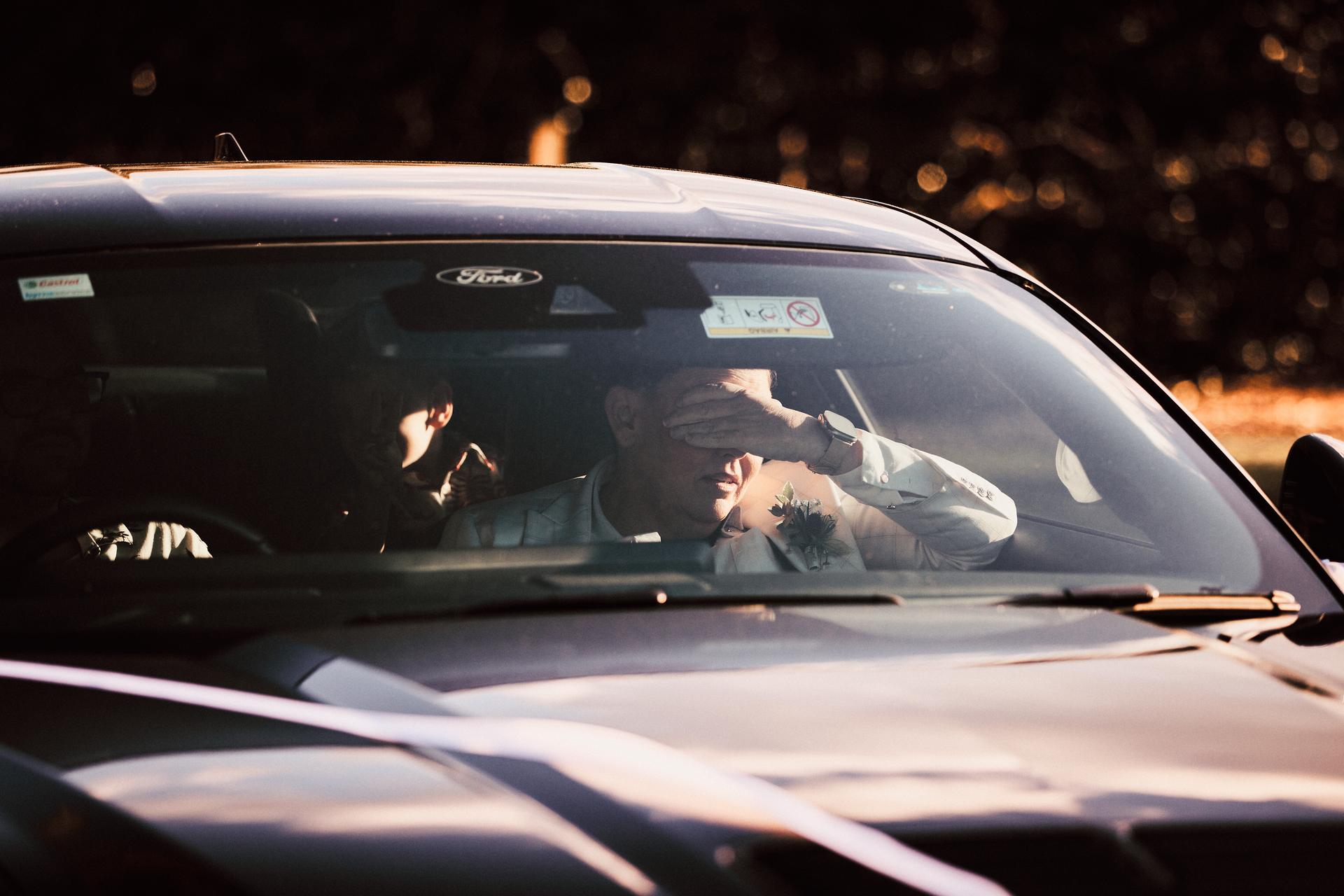 A man wearing a boutonniere sits in the front passenger seat of a Ford car, shielding his eyes from the sun with his hand, while another person is partially visible in the back seat.