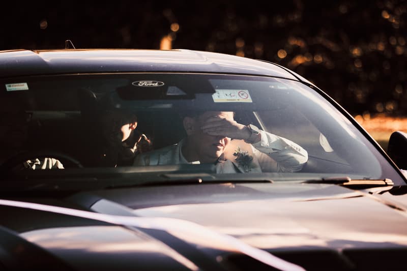 A man wearing a boutonniere sits in the front passenger seat of a Ford car, shielding his eyes from the sun with his hand, while another person is partially visible in the back seat.