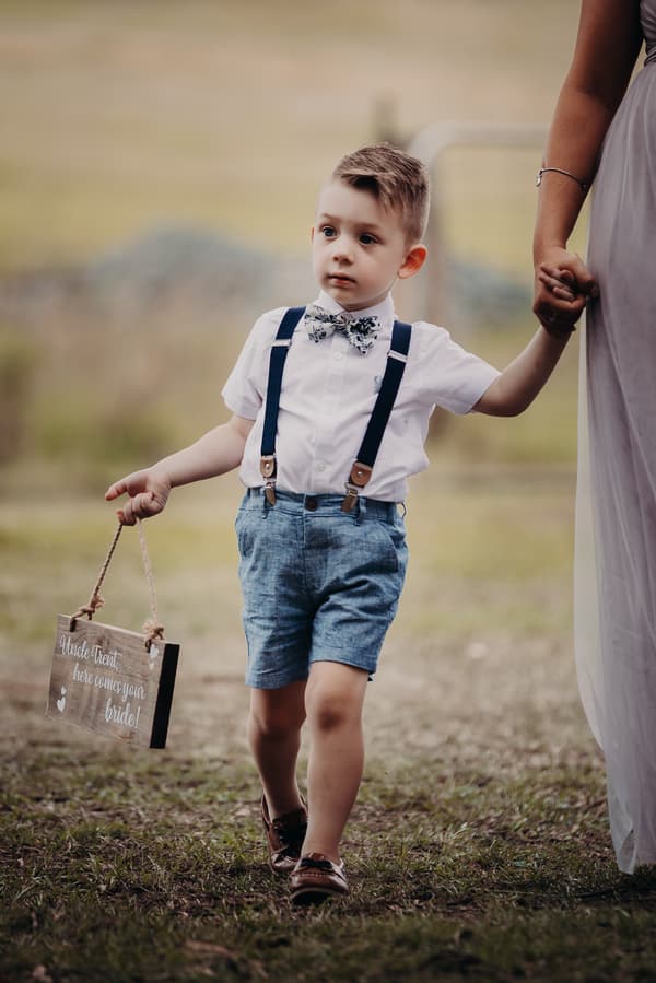 A young boy dressed in a white shirt, blue shorts, suspenders, and a bow tie holds a wooden sign that reads 'Uncle Trent, here comes your bride!' while holding the hand of a woman in a lavender dress at Ocean View Estates.
