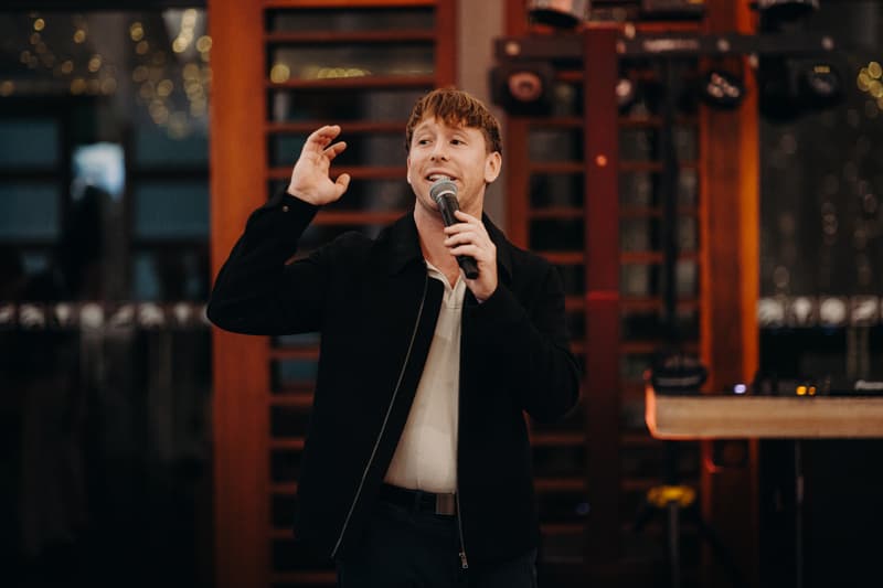 A man in a black jacket and white shirt holds a microphone and speaks on the reception stage at The Tides — The Pandanus Room.