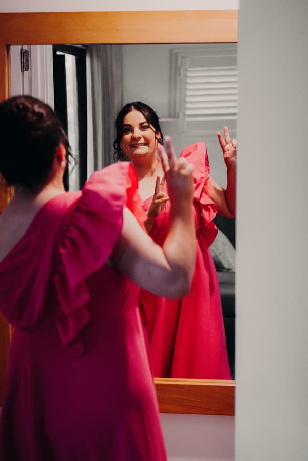 A bridesmaid in a bright pink dress makes a peace sign while looking at herself in a mirror at Sandstone Point Hotel.