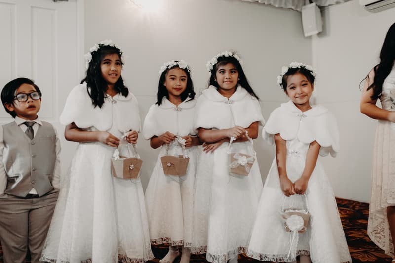 Four flower girls in white dresses and white shawls holding flower baskets stand next to a young boy in a grey suit at Royal on the Park.