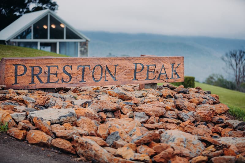 Wooden sign reading 'PRESTON PEAK' mounted on a bed of rocks with a building and misty hills in the background at Preston Peak Winery Functions Centre.