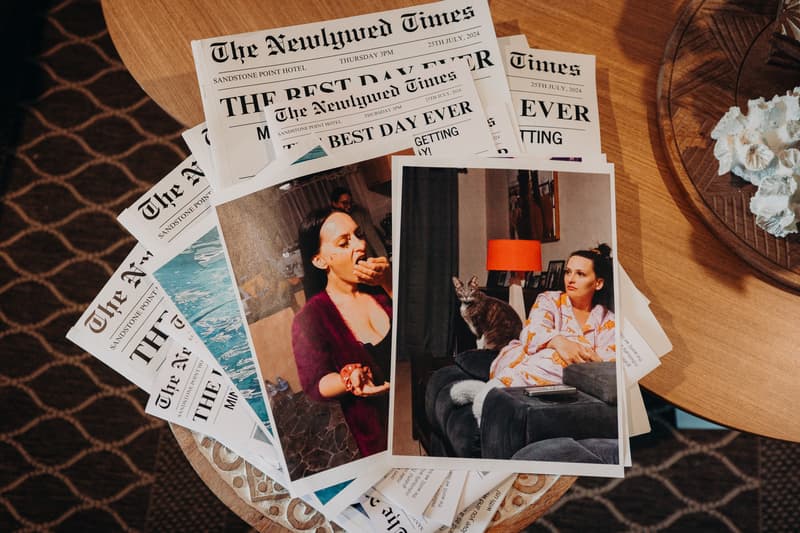 A stack of wedding-themed newspapers titled 'The Newlywed Times' from Sandstone Point Hotel dated 25th July 2024, with two printed photographs on top showing a woman eating and a woman sitting on a couch with a cat.