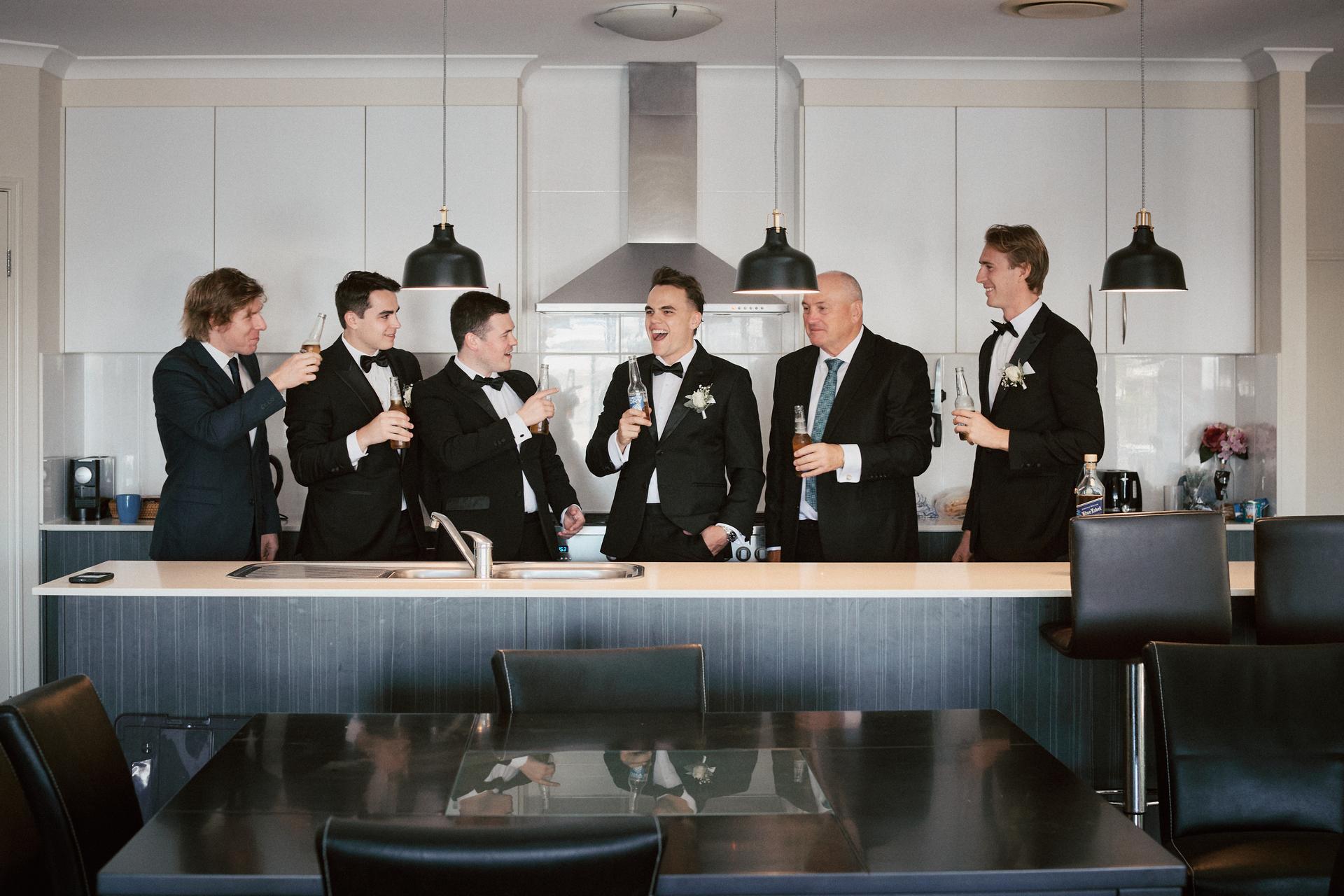 James and five groomsmen stand behind a kitchen island at Sandstone Point Hotel, holding drinks and laughing together.