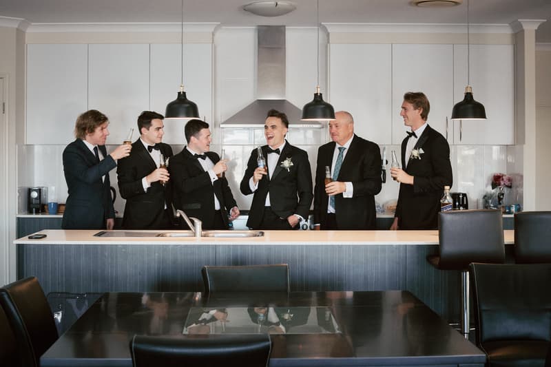 James and five groomsmen stand behind a kitchen island at Sandstone Point Hotel, holding drinks and laughing together.