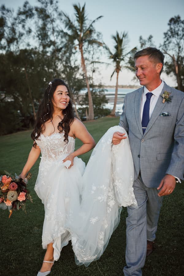 The bride Jheng and the groom Dane walk on grass at Sandstone Point Hotel. Jheng holds a bouquet and lifts her wedding dress train, while Dane holds part of the train and wears a light grey suit with a blue tie and boutonniere.