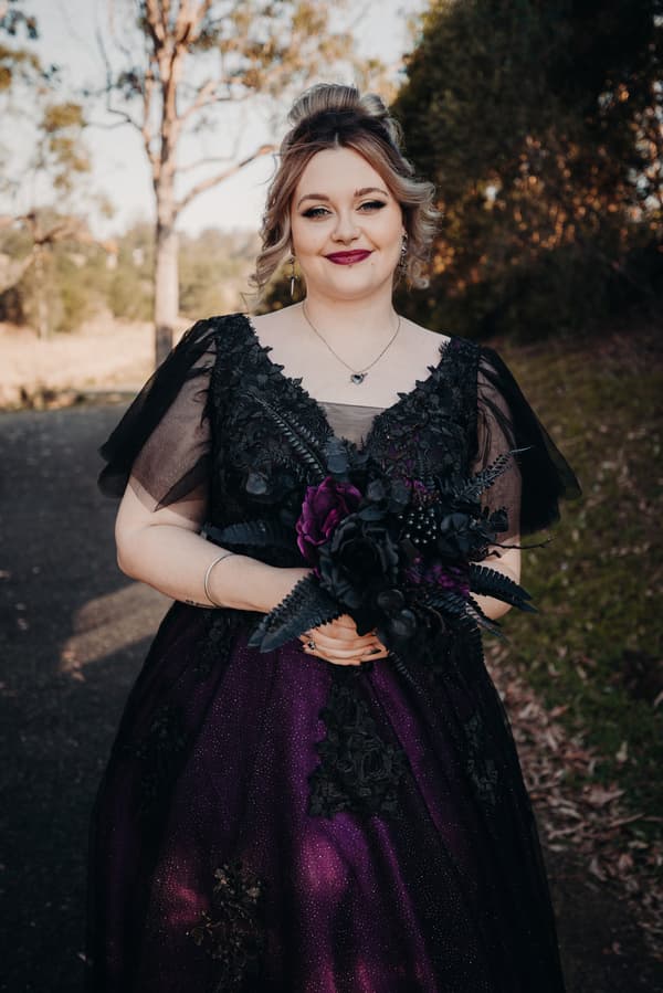 The bride stands outdoors at Ocean View Estates — On The Lake, wearing a dark purple and black gown and holding a bouquet of black and purple flowers.