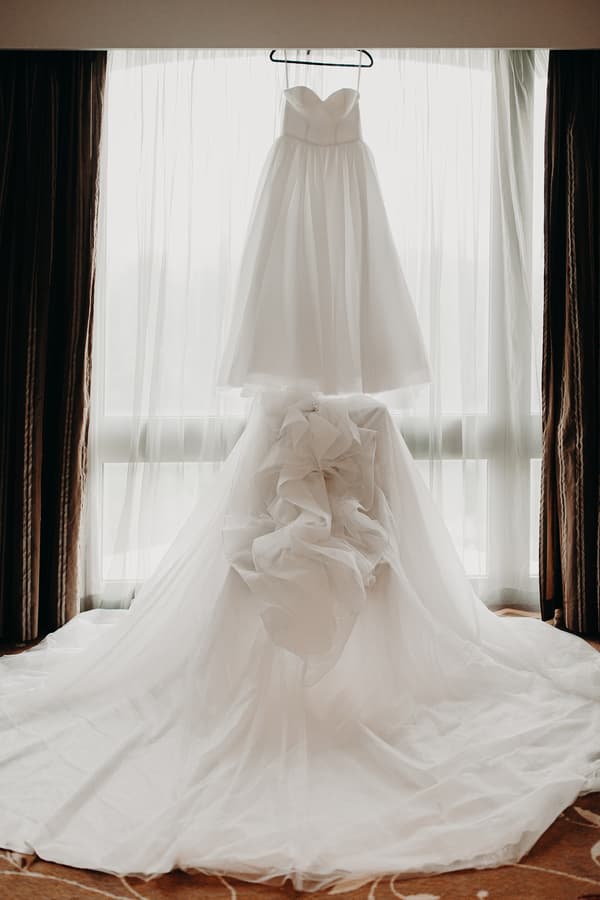 The bride's wedding dress is displayed hanging in front of a window with sheer curtains at Royal on the Park.