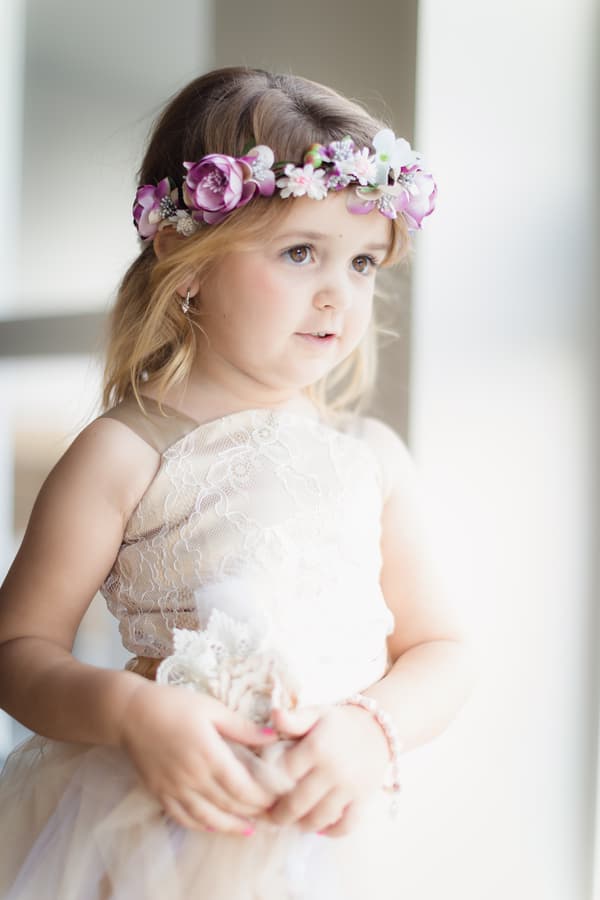 A young flower girl wearing a lace dress and a floral headband stands indoors near a window at Novotel Sunshine Coast Resort.