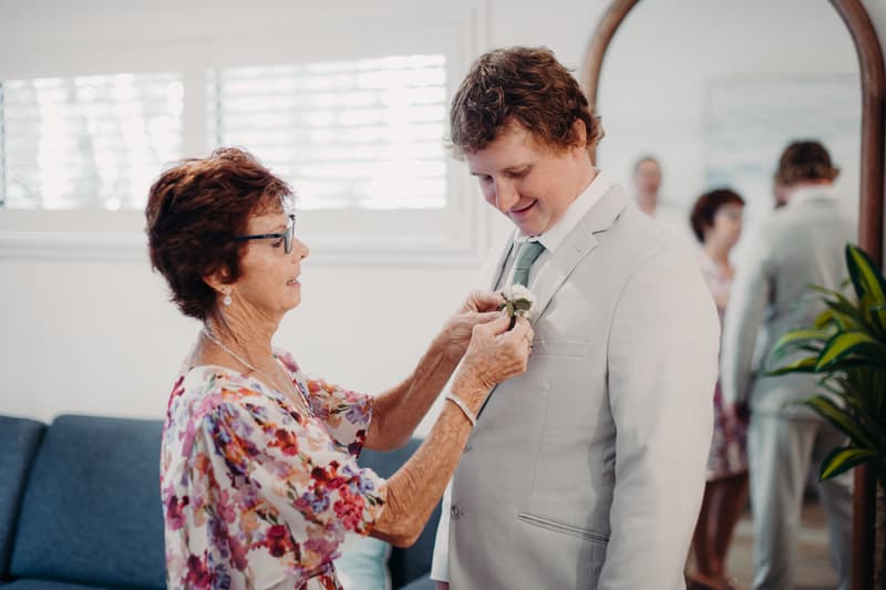 The groom Brodie has a boutonniere pinned on his light grey suit jacket by an older woman, likely his mother, inside a room at Sandstone Point Hotel.