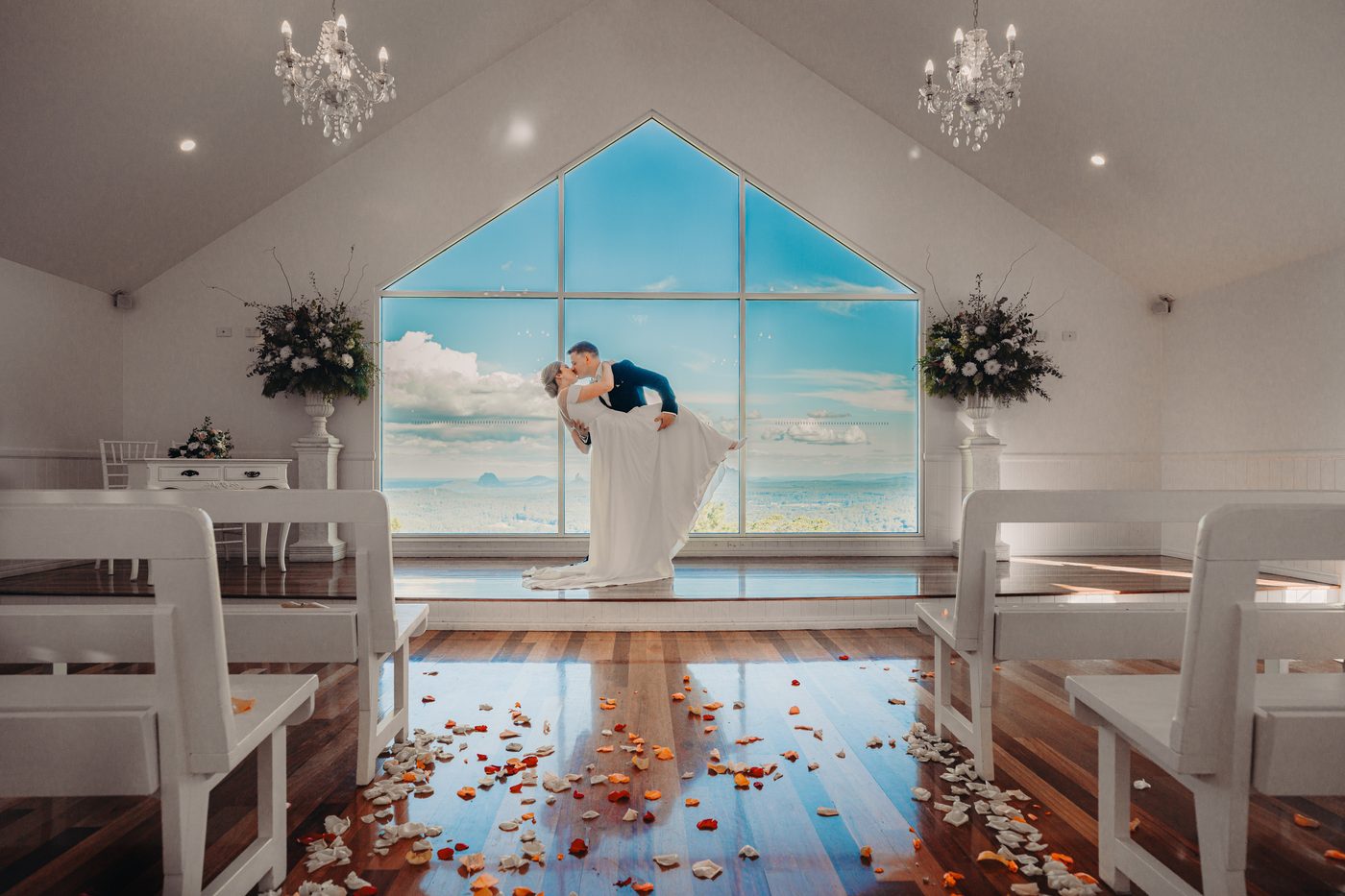Courtney and Liam pose for a couple portrait inside, with Liam dipping Courtney in front of a large triangular window showing a scenic sky view. The chapel has white benches, wooden floors scattered with rose petals, chandeliers, and floral arrangements on pedestals. Elopement photography packages Queensland by She Said Yes.