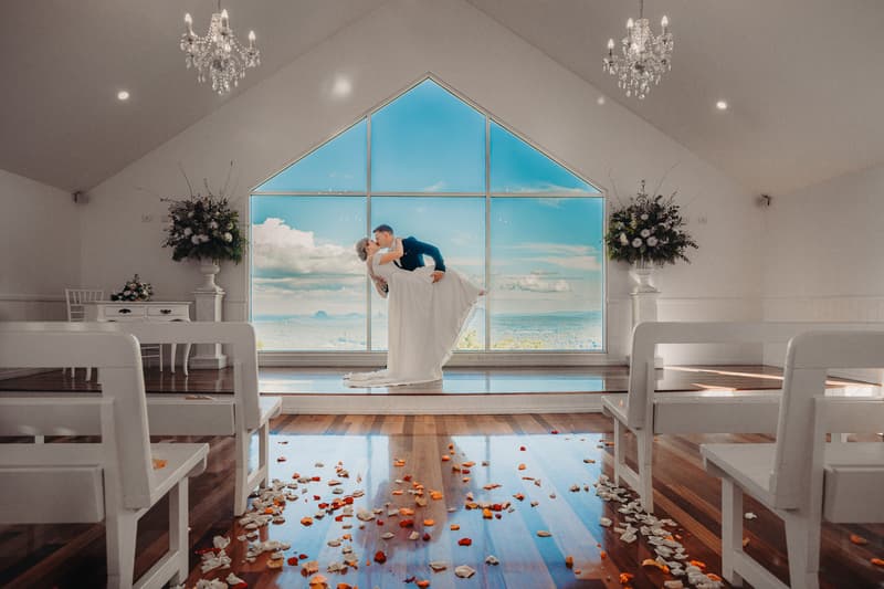 Courtney and Liam pose for a couple portrait inside Tiffany's Maleny chapel, with Liam dipping Courtney in front of a large triangular window showing a scenic sky view. The chapel has white benches, wooden floors scattered with rose petals, chandeliers, and floral arrangements on pedestals.