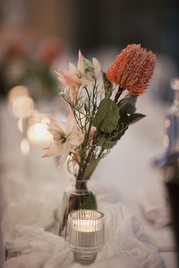 Close-up of a floral centerpiece with pink and orange flowers in a glass vase alongside a lit candle on a white draped table at Sandstone Point Hotel — The Pumicestone Room.