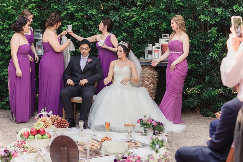 Bride Maryam and groom Pasha seated outdoors surrounded by bridesmaids in purple dresses at Hillstone St Lucia — The Quartyard, with a low table set with food and flowers in front of them.