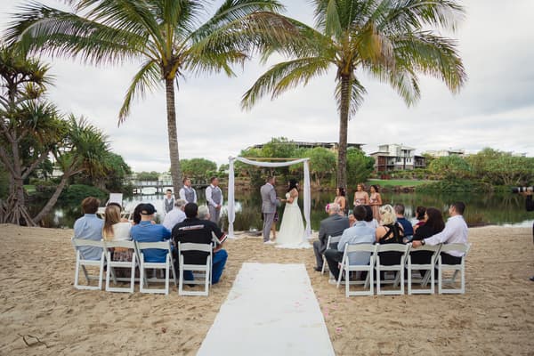 Kelly and Shane stand under a white fabric-draped arch exchanging vows at their wedding ceremony on a sandy area by a water body at Novotel Sunshine Coast Resort, with bridesmaids, groomsmen, and seated guests watching.