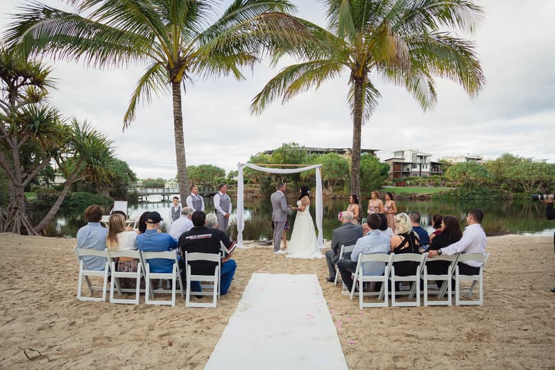Kelly and Shane stand under a white fabric-draped arch exchanging vows at their wedding ceremony on a sandy area by a water body at Novotel Sunshine Coast Resort, with bridesmaids, groomsmen, and seated guests watching.