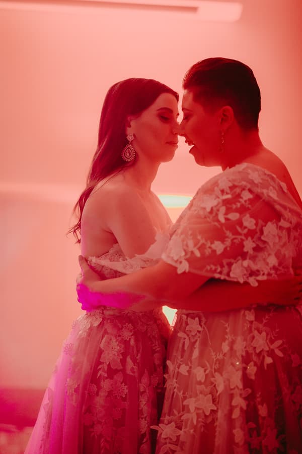 Two brides in floral lace wedding dresses embrace closely indoors under pink lighting.