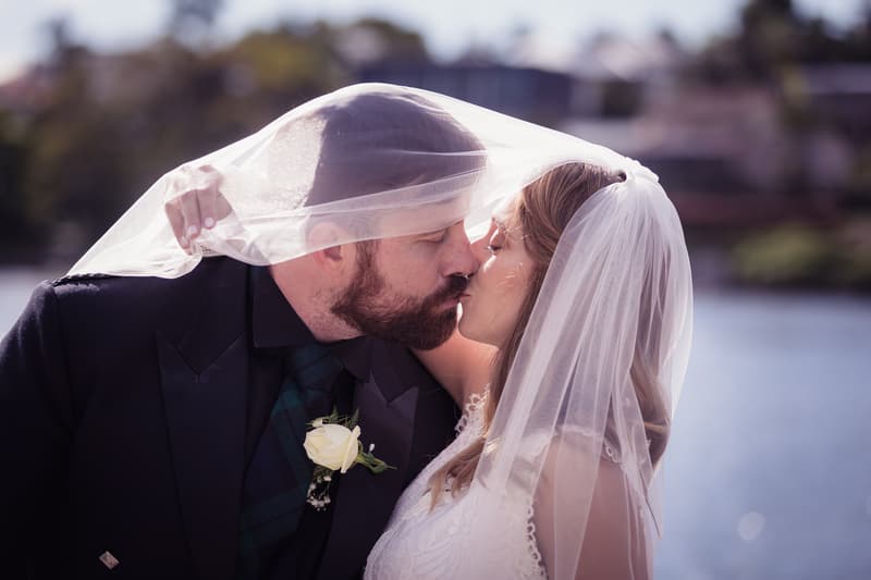 Francesca and Ben share a kiss with Francesca's veil draped over both of them at Toowong Rowing Club — The Malouf Room, with water and blurred greenery in the background.