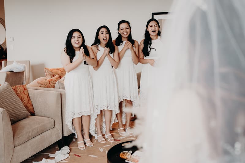 Four bridesmaids in matching white dresses stand indoors at Royal on the Park, reacting with surprise and joy to the bride who is partially visible in the foreground wearing a veil.