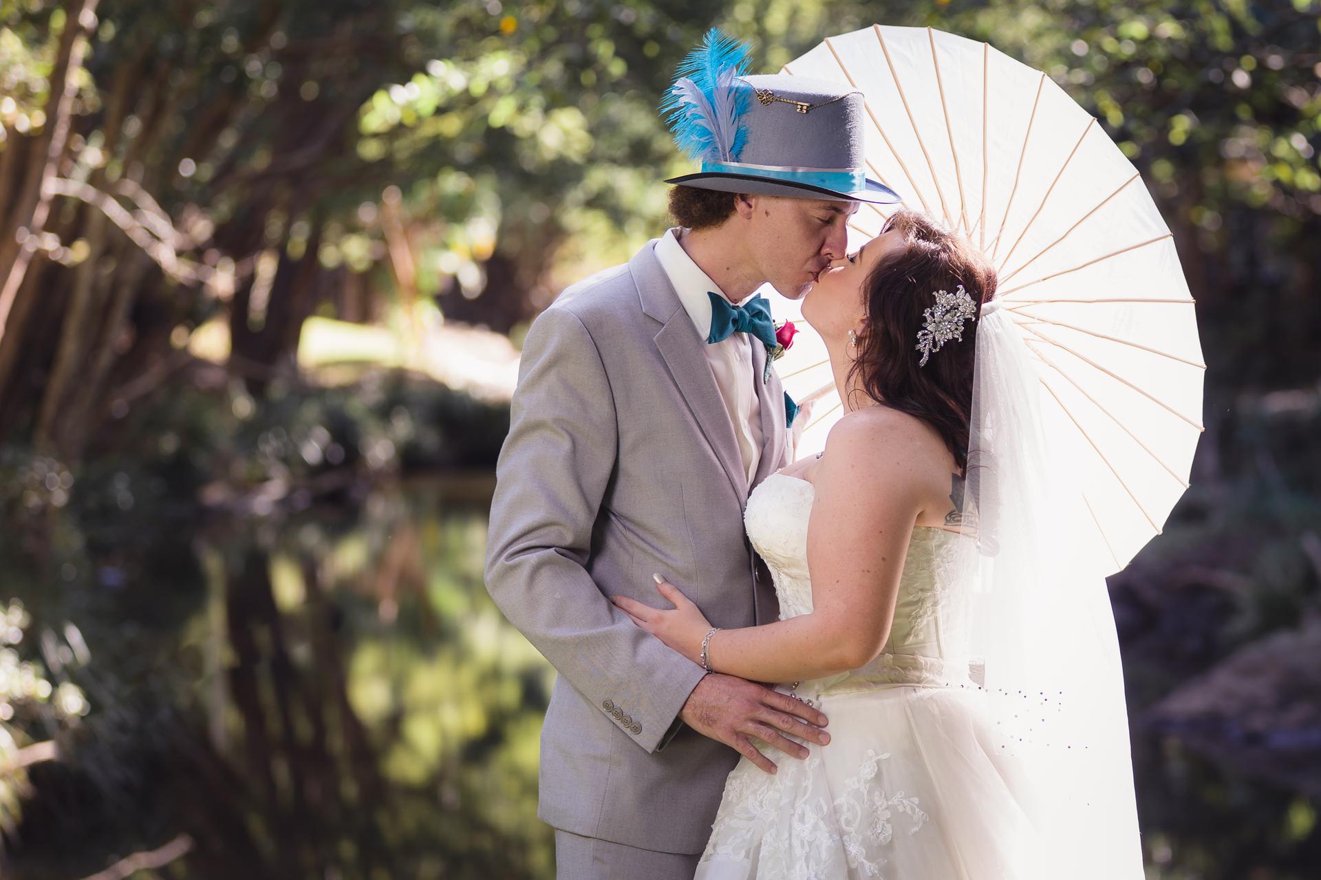 Bride Holly and groom Paul kiss outdoors near a pond at Kwila Lodge, with Holly holding a white parasol and Paul wearing a grey suit with a blue feathered hat.
