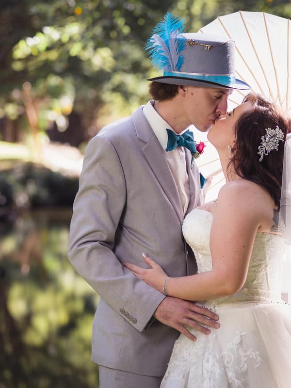 Bride Holly and groom Paul kiss outdoors near a pond at Kwila Lodge, with Holly holding a white parasol and Paul wearing a grey suit with a blue feathered hat.