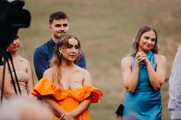 Guests stand outdoors during the wedding ceremony, including a woman in an orange off-shoulder dress and a woman in a blue sleeveless dress. Basic ceremony video by She Said Yes.