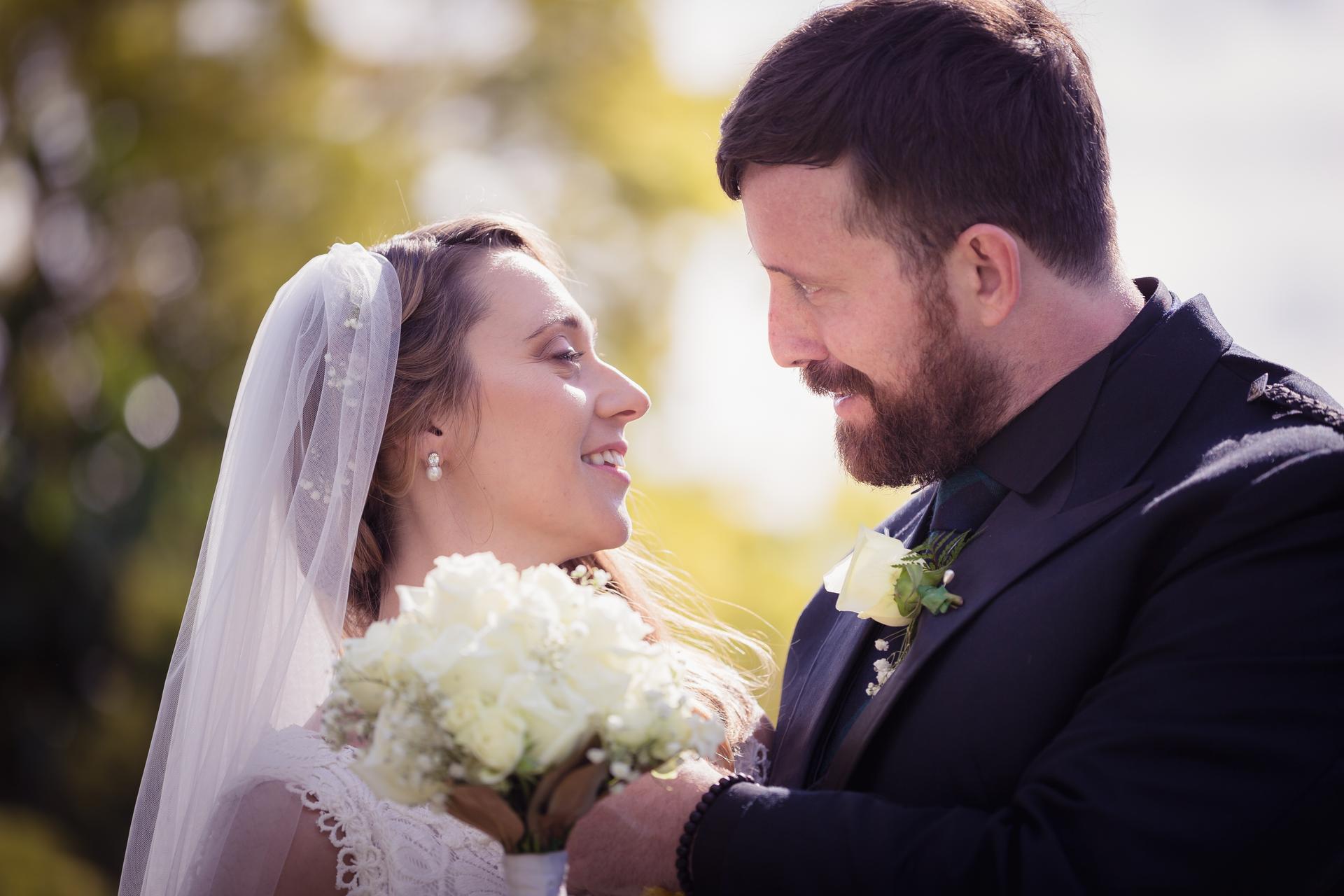Bride Francesca and groom Ben share a close moment outdoors, with Francesca holding a bouquet of white flowers and wearing a veil, and Ben dressed in a dark suit with a white rose boutonniere.