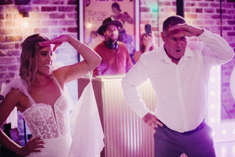The bride Krystal and a male guest dance together on the reception stage at Sandstone Point Hotel — Cellar, with a DJ in the background.