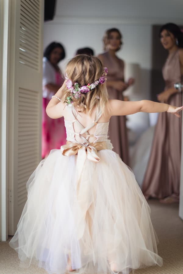 A young flower girl wearing a white dress with a large bow and a floral headband stands with her back to the camera, arms outstretched, facing three bridesmaids in matching mauve dresses inside a room at Novotel Sunshine Coast Resort.