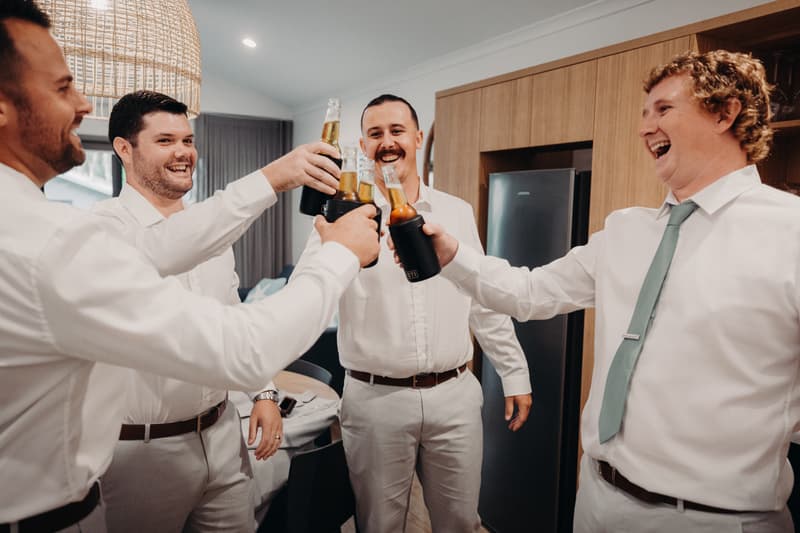 The groom and three groomsmen toast with bottled drinks inside a room at Sandstone Point Hotel.