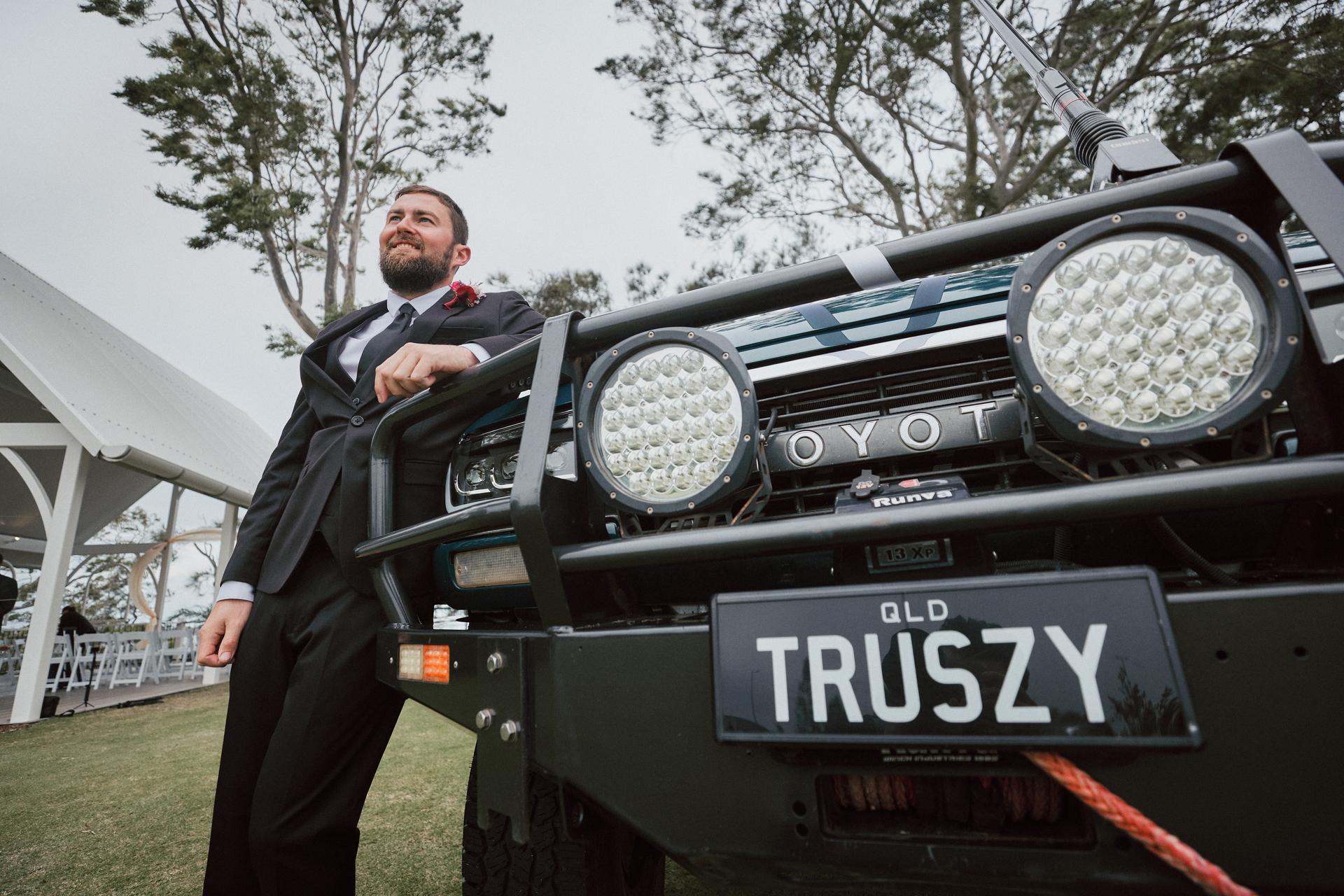 The groom in a black suit leans against the front of a black Toyota vehicle with a Queensland license plate reading 'TRUSZY' at Sandstone Point Hotel — The Pavilion.