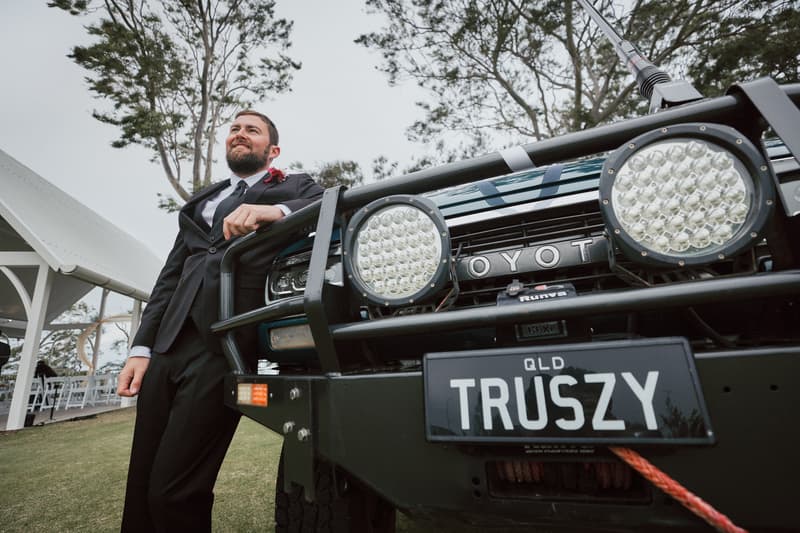 The groom in a black suit leans against the front of a black Toyota vehicle with a Queensland license plate reading 'TRUSZY' at Sandstone Point Hotel — The Pavilion.