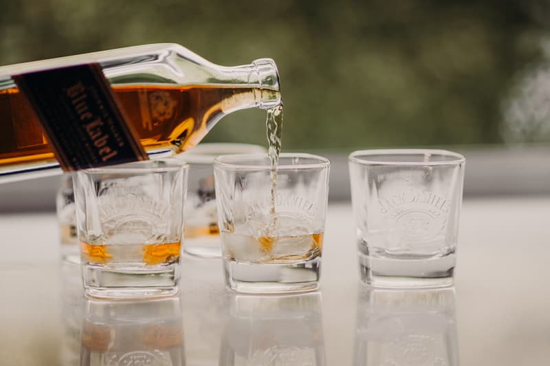 Whiskey is being poured from a bottle of Jack Daniel's into a glass on a reflective surface with three other empty or partially filled Jack Daniel's glasses nearby.